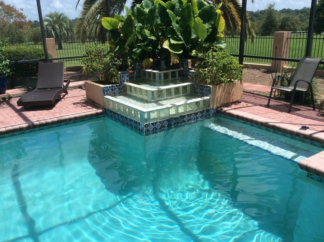 A turquoise backyard swimming pool with a tiered fountain planter and two lounge chairs on a brick patio.