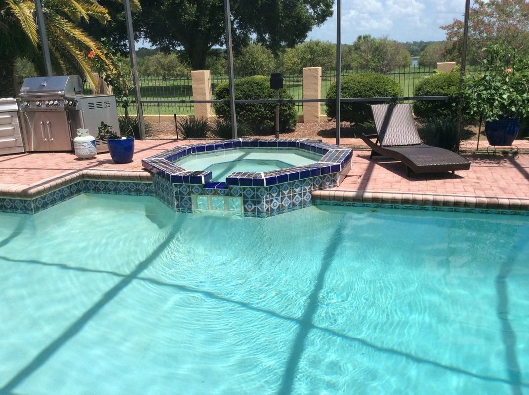 A tiled hot tub spilling into a swimming pool on a brick patio with a lounge chair, grill, and outdoor screen enclosure.