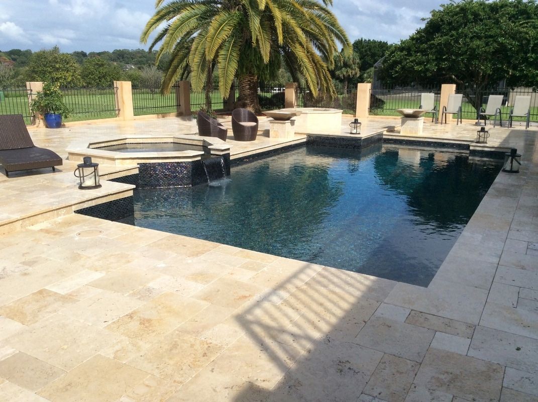 A rectangular swimming pool with a hot tub and dark stone water features, set on a beige stone patio with palm trees.