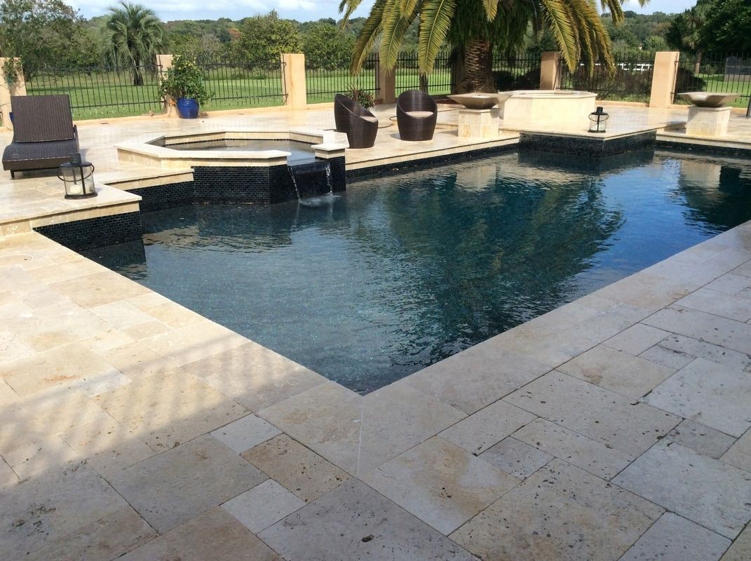 A rectangular pool with a dark tiled interior and an attached hot tub, surrounded by beige stone pavers on a sunny day.
