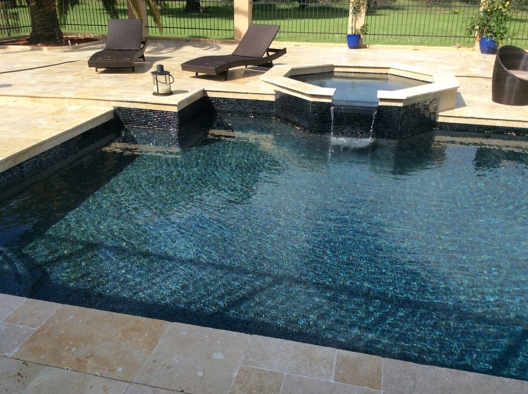 A dark-tiled swimming pool with a stone spillover spa, surrounded by lounge chairs on a light-colored stone patio.