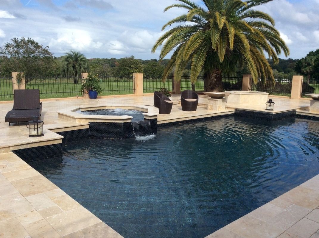 A swimming pool with an attached hot tub, surrounded by a stone patio, lounge chairs, and a large palm tree.