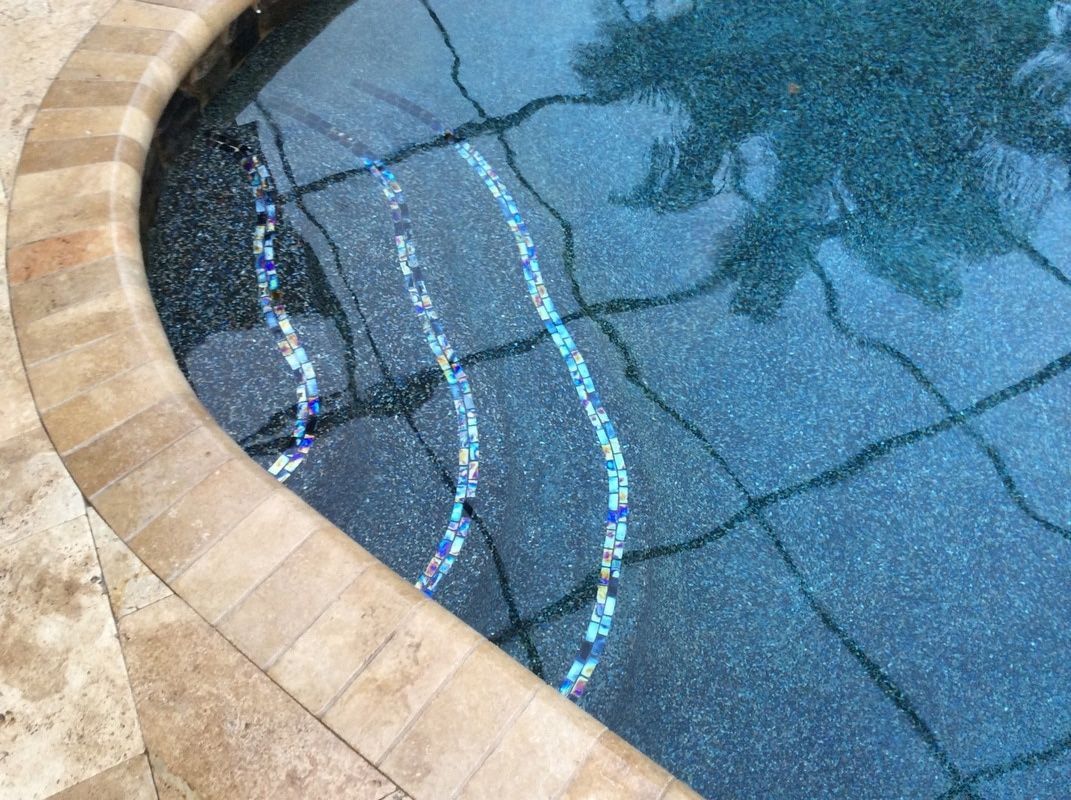A curved pool edge with tan stone coping leading into deep blue water with tiled steps and light-refraction patterns.