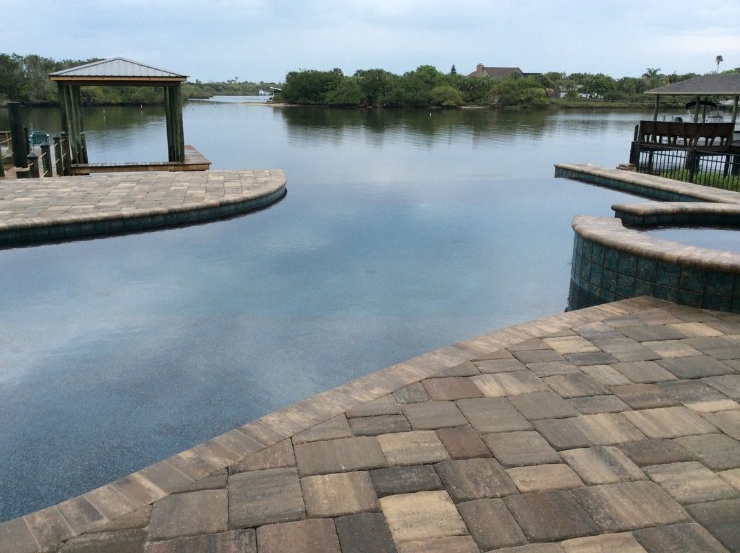 A stone-paved patio borders an infinity pool overlooking a calm lake and distant trees under an overcast sky.