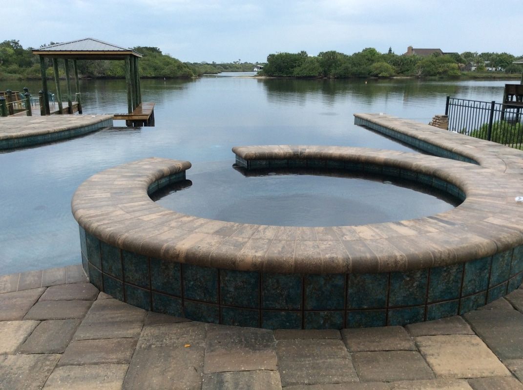 A circular, tiled hot tub with a stone coping edge sits on a patio overlooking a calm lake and distant treeline.