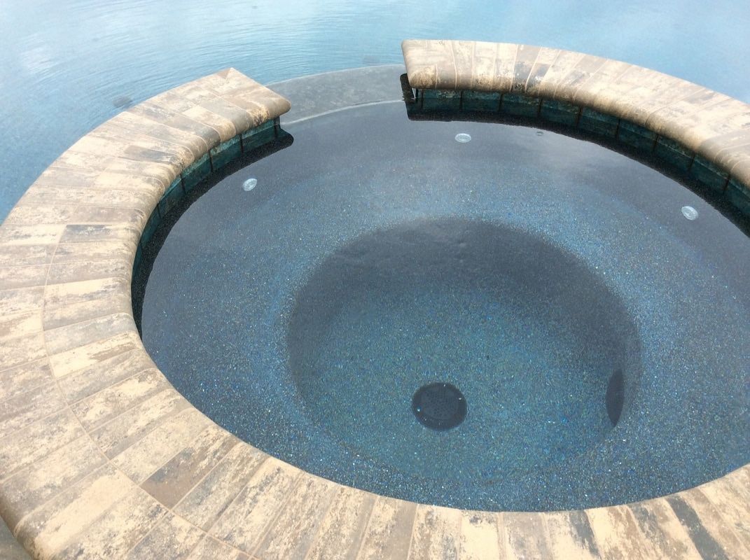 A circular stone hot tub spa with dark, textured interior tiles, partially submerged in a larger swimming pool.