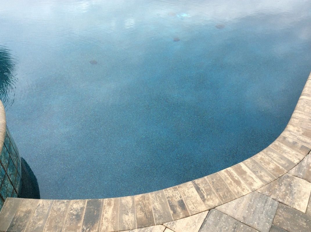 A high-angle view of a swimming pool with dark blue water and a curved stone paver border.
