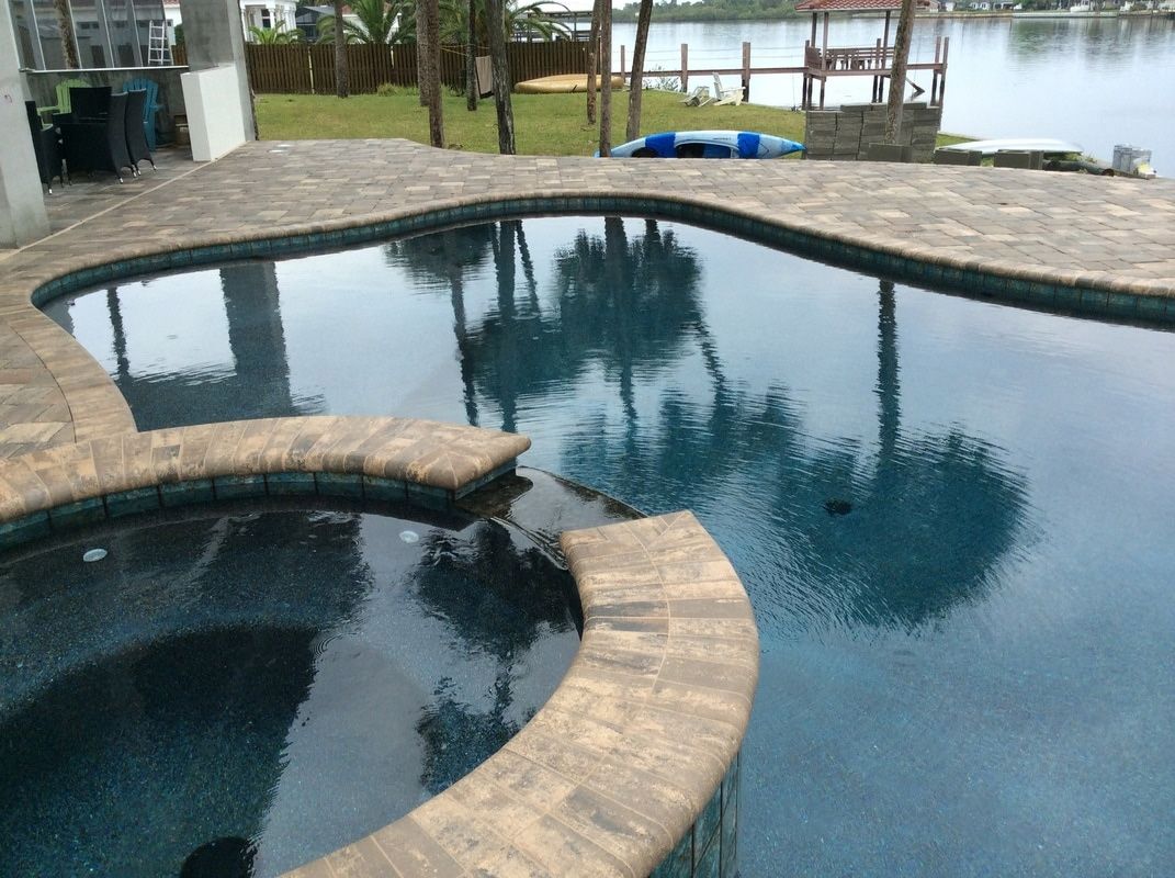 An infinity-edge swimming pool and attached hot tub with pavers, overlooking a calm lake with a wooden dock.