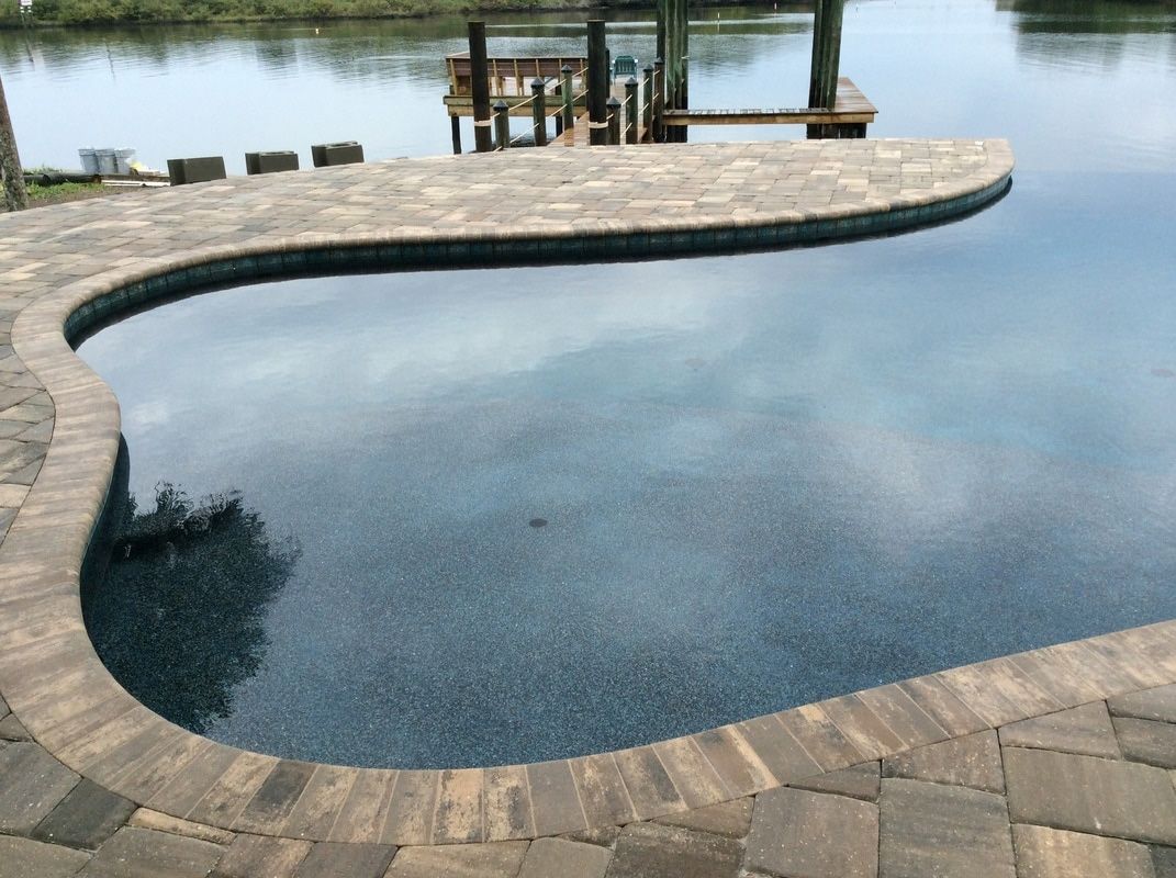 A curved swimming pool with dark blue water and stone paver decking, situated next to a canal and a wooden dock.