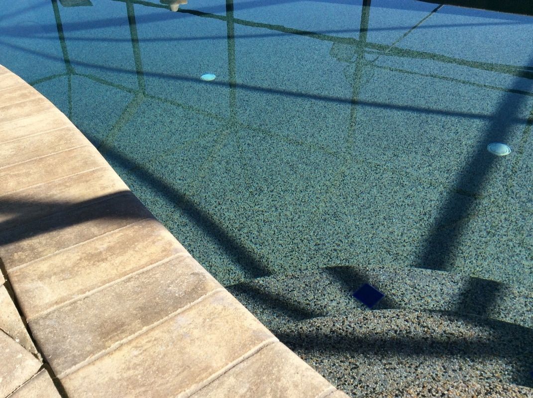 A view of pool steps submerged in clear water next to a textured concrete pool deck, seen through a pool screen enclosure.