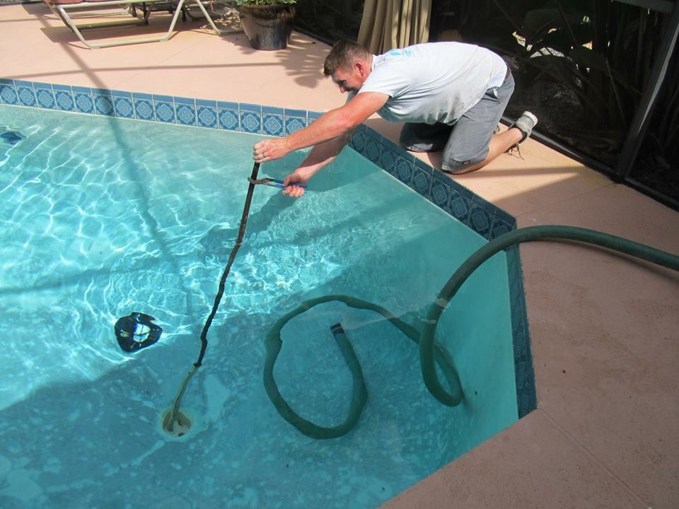 A person crouched by a pool deck uses long-handled shears to cut a dark hose submerged in the swimming pool water.