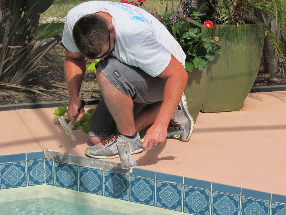 A person kneeling by a swimming pool uses a mallet to secure trim along the edge of the blue-patterned tile waterline.