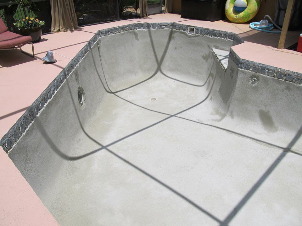 An empty, light-colored concrete swimming pool with a tile border, set within a patio area.