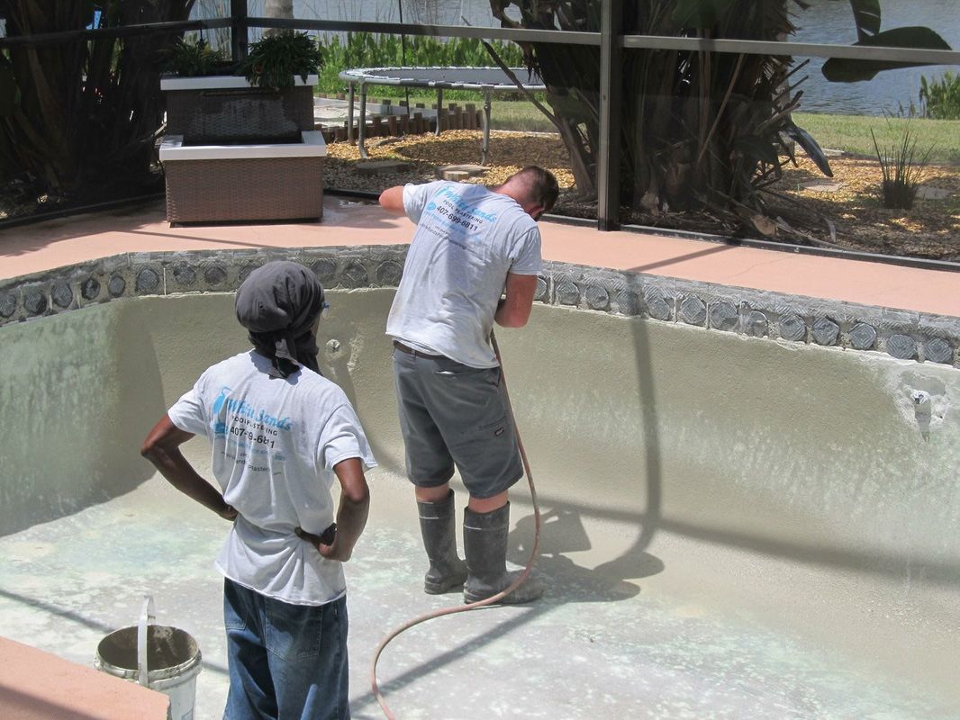 Two people in a drained backyard swimming pool, with one person using a power tool to smooth the pool's interior wall.