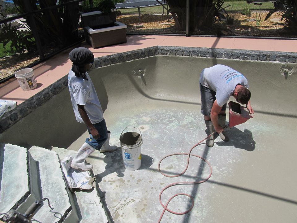 Two people in protective footwear applying plaster to the interior of an empty, light-colored swimming pool.