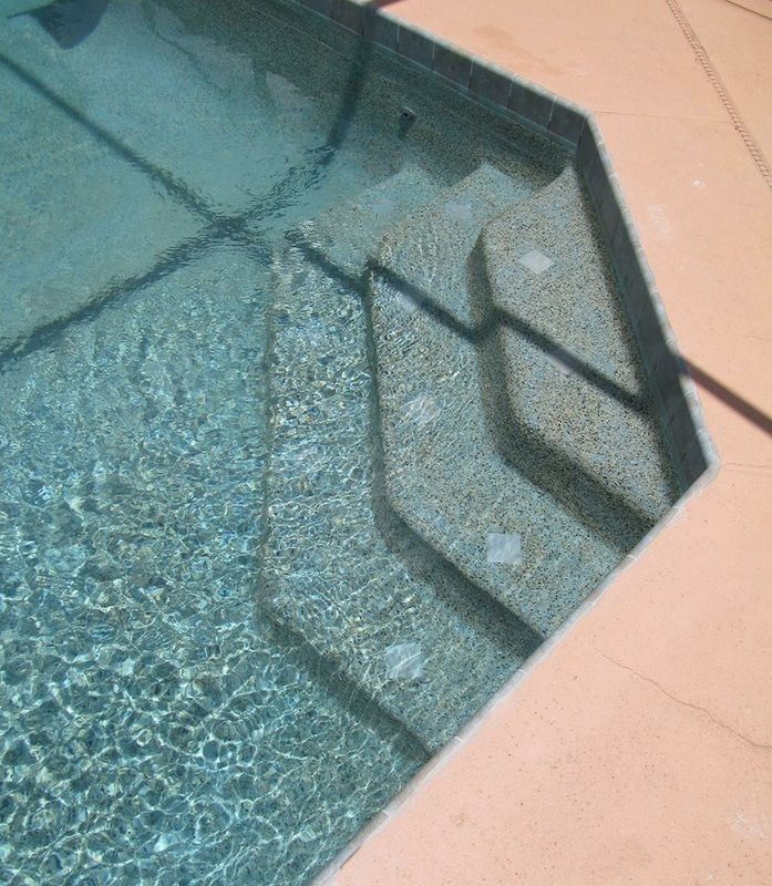 Steps leading into a swimming pool with light-colored stone aggregate finish and beige concrete decking.