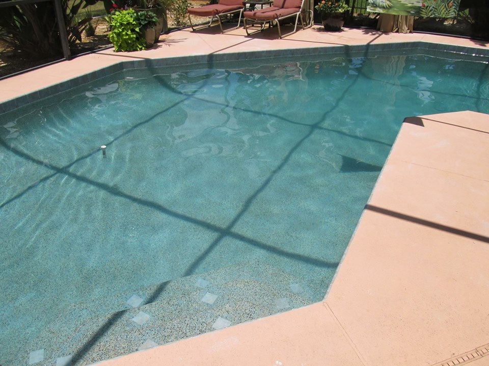 A clear, blue rectangular swimming pool with steps, bordered by a light-colored paved deck and patio furniture.