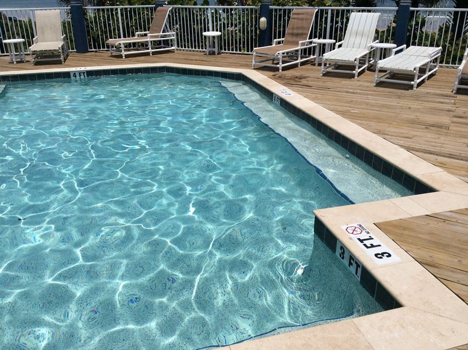A turquoise swimming pool surrounded by a light-colored wooden deck with several white lounge chairs and small side tables.