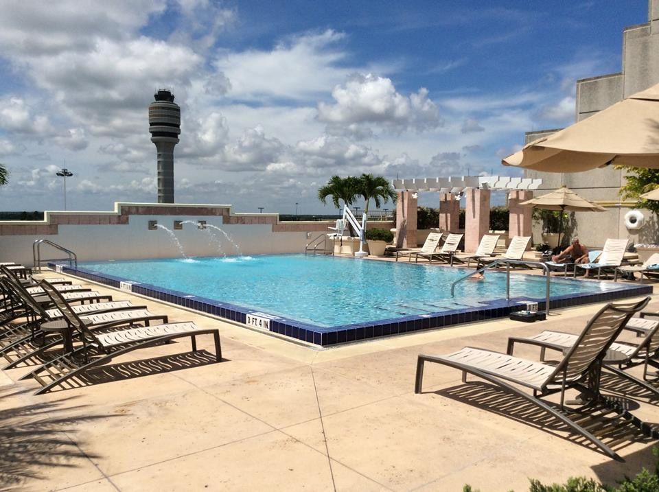 An outdoor hotel pool with lounge chairs, umbrellas, and a prominent airport control tower in the background.