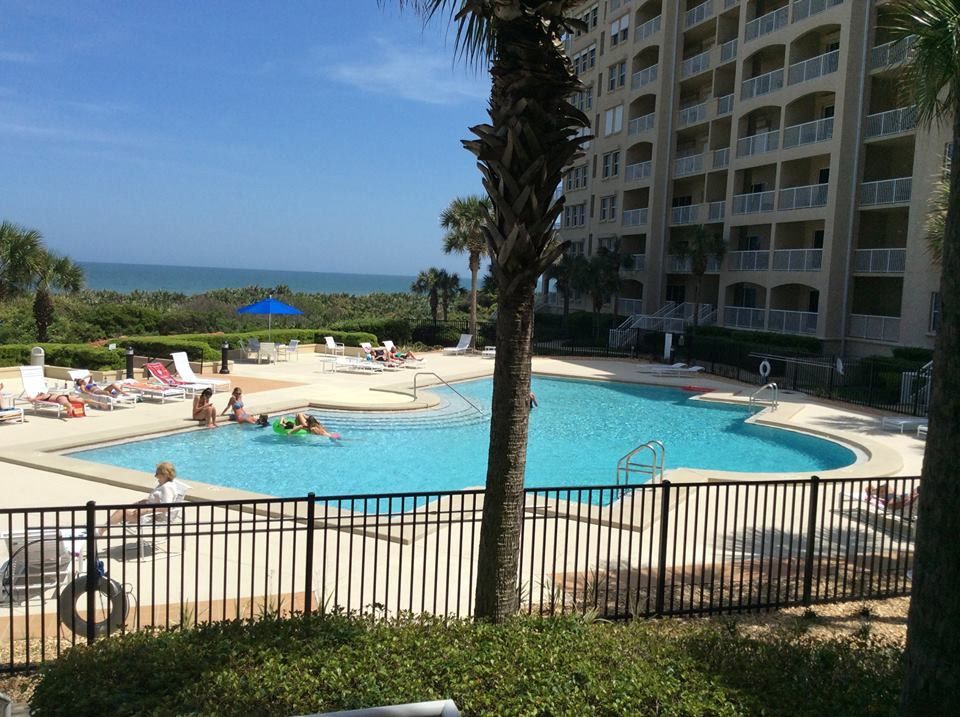 A resort swimming pool next to a multistory building overlooking a beach with ocean views on a sunny day.