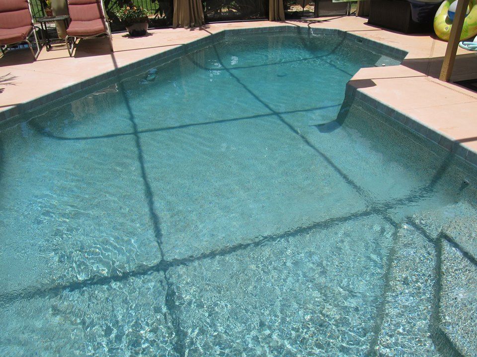 A clear, irregularly shaped backyard swimming pool with light-colored steps and patio furniture on the deck.