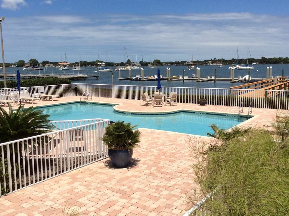 A swimming pool on a patio overlooking a bay filled with sailboats and a wooden pier under a sunny blue sky.