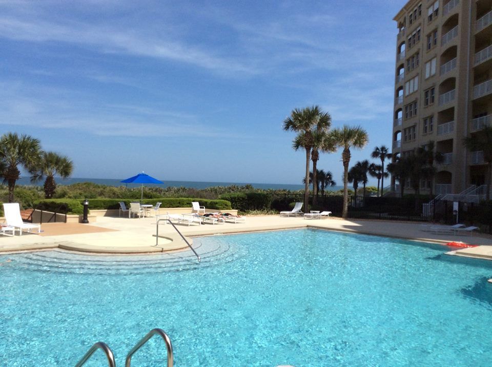 Outdoor swimming pool next to a tall building and palm trees, with a blue ocean horizon in the distance.