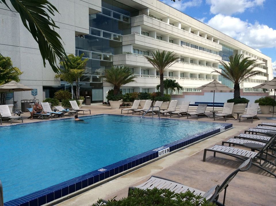 Swimming pool in front of a multi-story hotel with lounge chairs on the patio under a blue sky with scattered clouds.