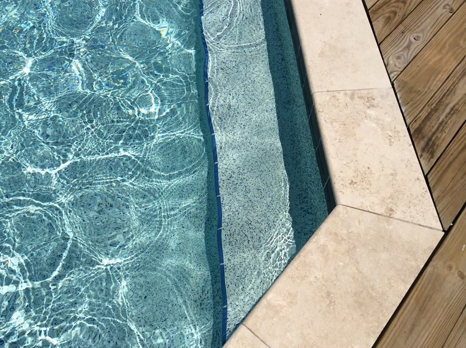 A corner of a swimming pool with light-colored stone coping and wooden decking bordering the water.