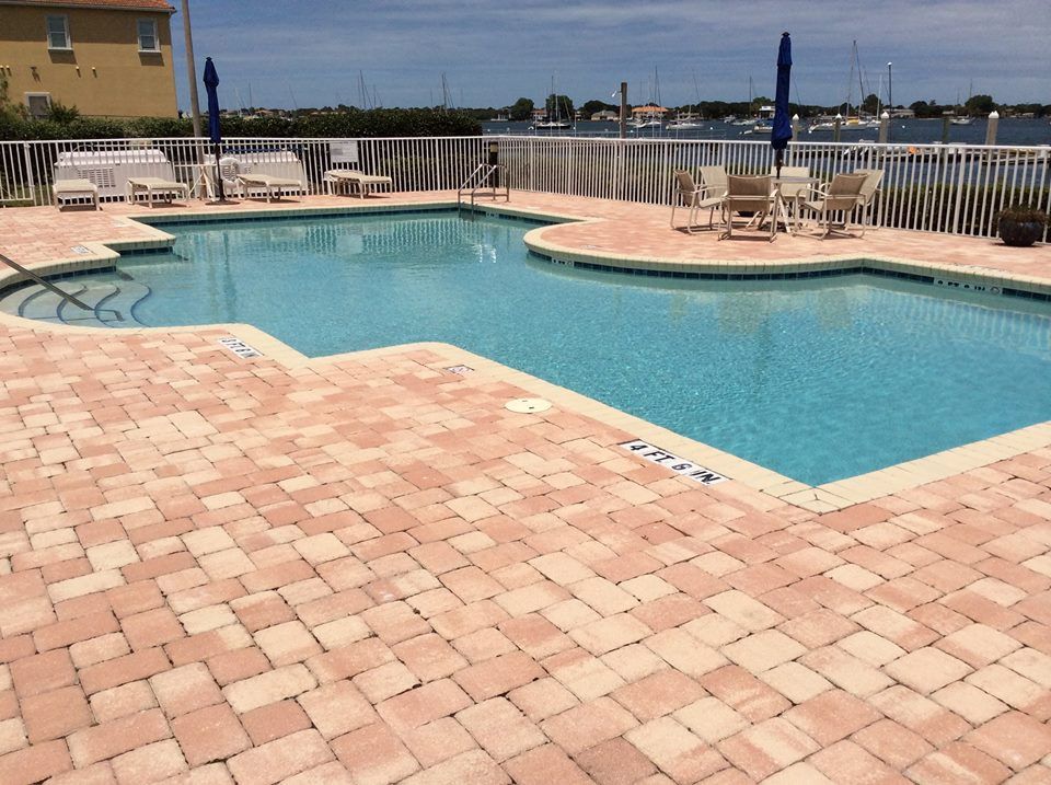 A light-colored brick pool deck surrounds a bright blue swimming pool overlooking a harbor with docked boats.