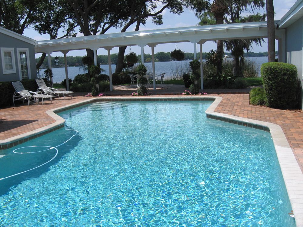 An outdoor swimming pool with blue water, surrounded by a brick patio, lounge chairs, and a covered walkway near a lake.