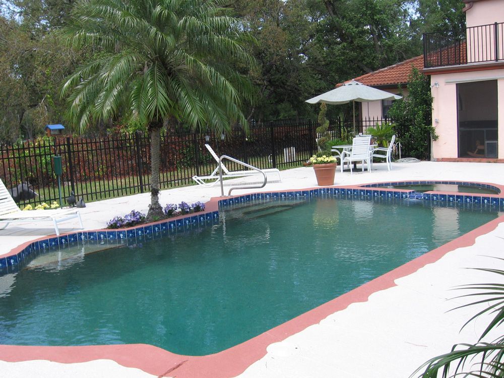 An outdoor swimming pool with a spa, a palm tree, and patio furniture on a paved deck next to a residential house.