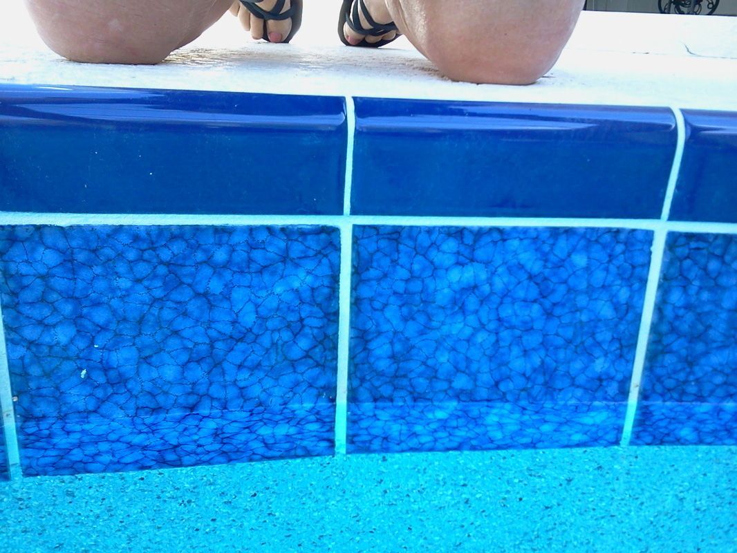 A person sits on the edge of a swimming pool with dark blue, textured tiles visible above the water surface.