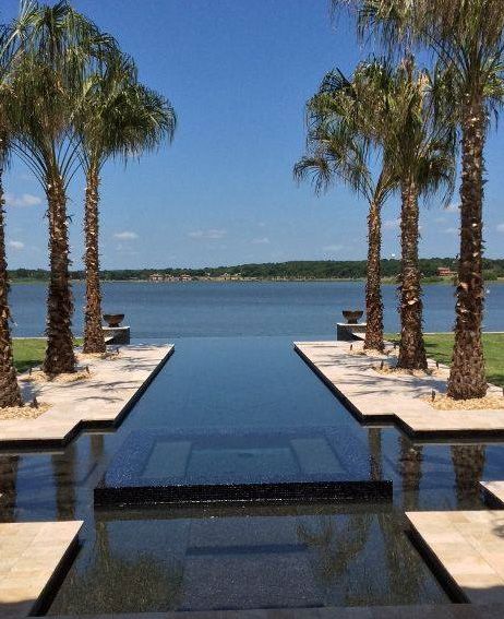 An infinity pool overlooks a calm lake, framed by tall palm trees under a bright blue sky.