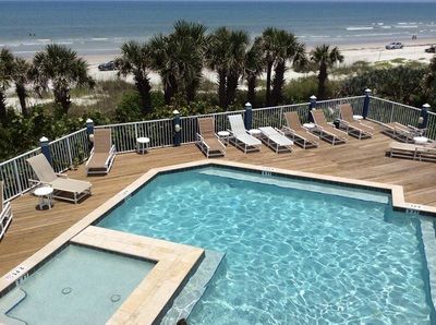 Swimming pool and hot tub on a wooden deck overlooking a beach with palm trees and the ocean under a clear blue sky.