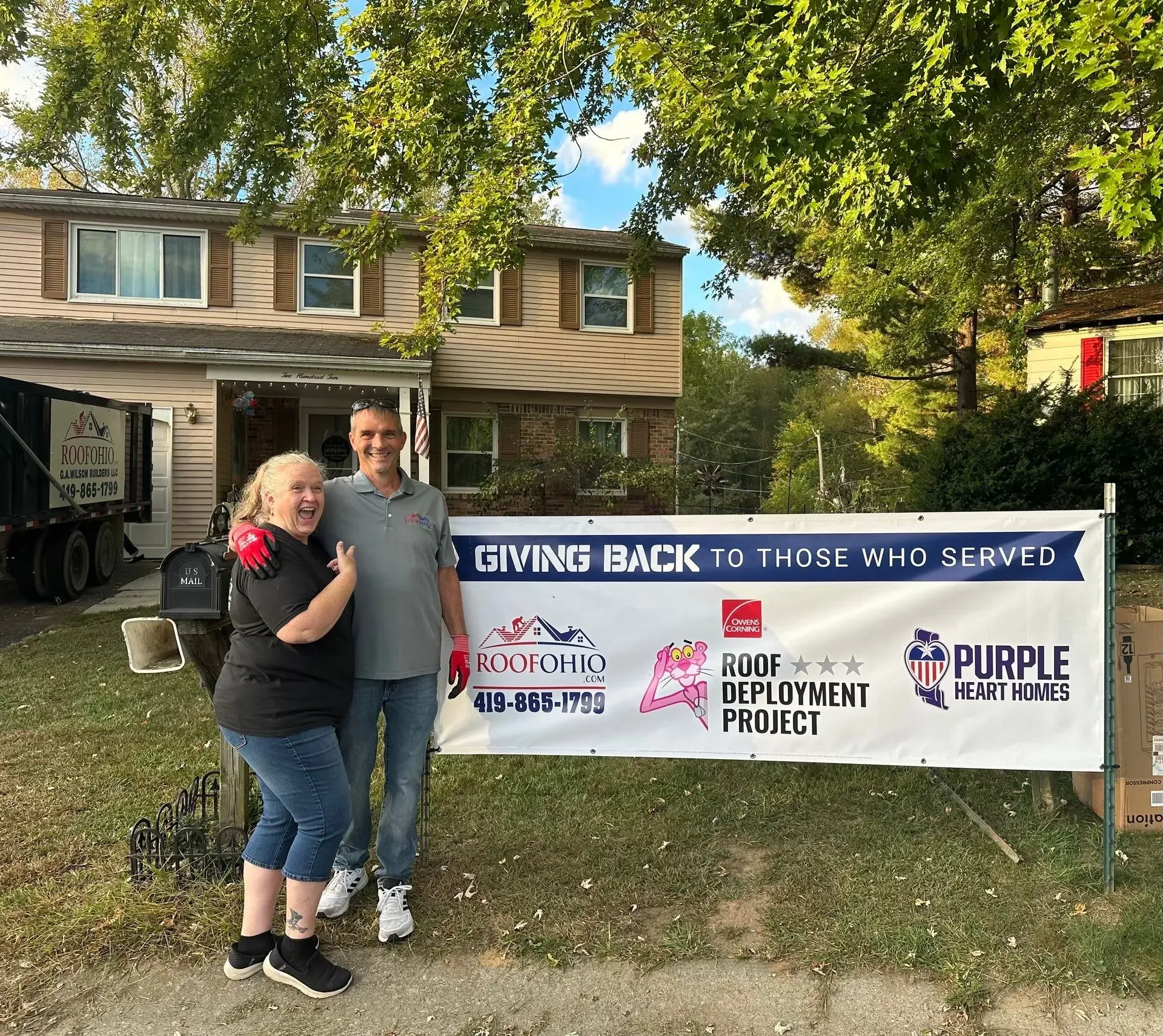 Three people stand in front of a house, with a banner that says,