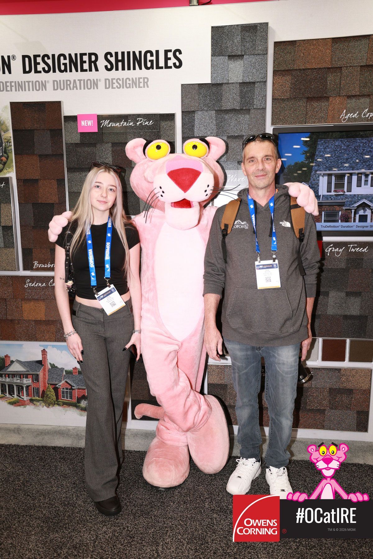 Two people posing with a pink mascot at a trade show booth backdrop