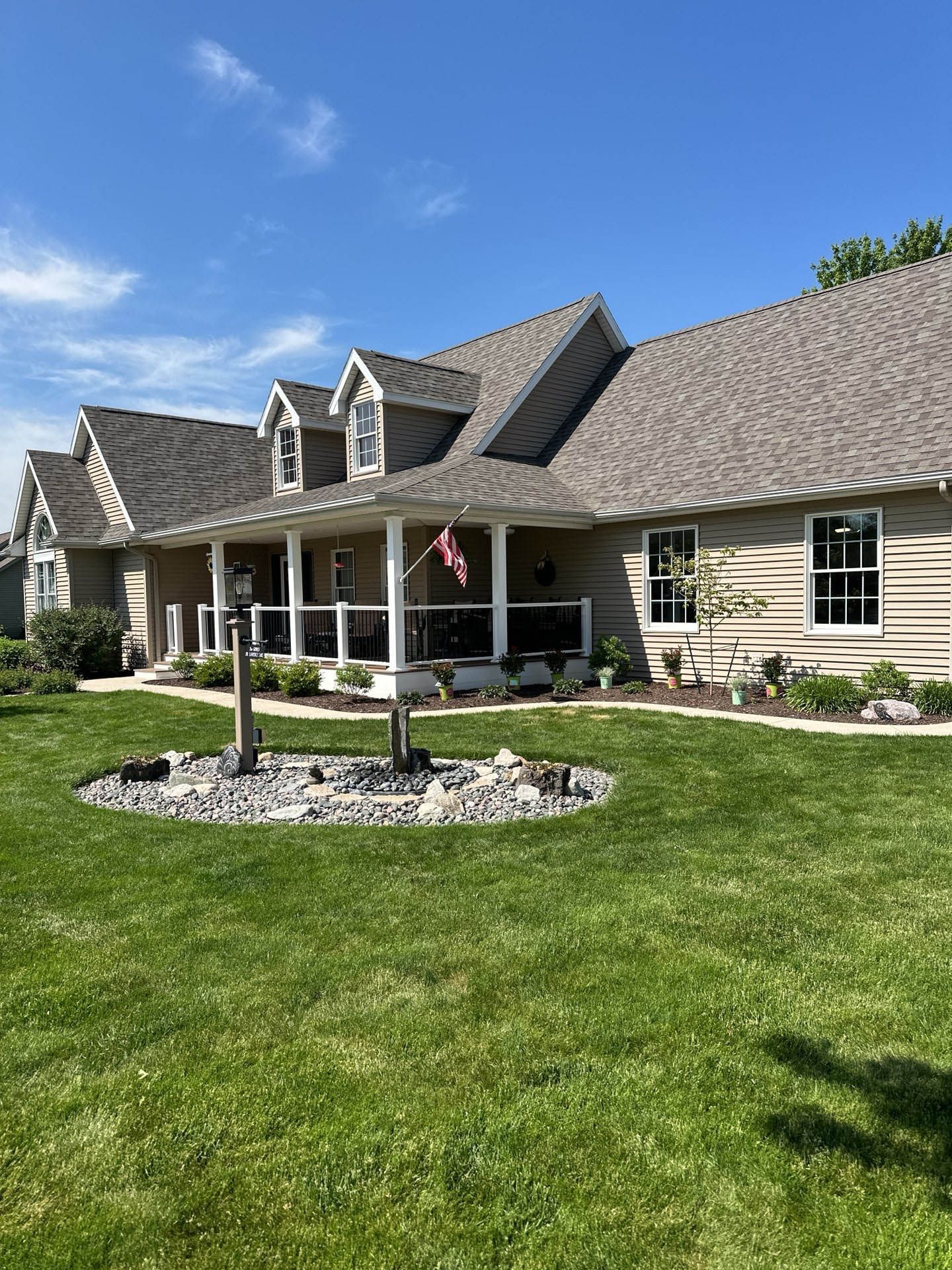 Single-story beige house with a gray roof, front porch, and large green lawn under a blue sky