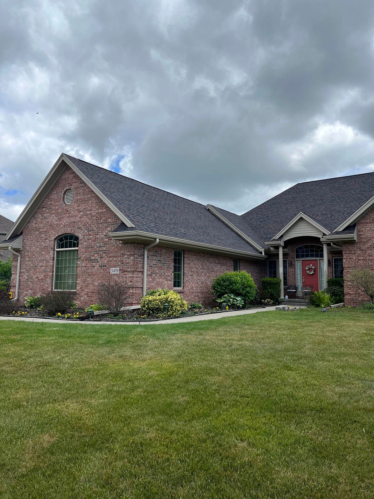 Brick suburban house with dark roof and red front door under cloudy sky, fronted by a green lawn