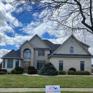 Suburban house with gray brick facade, green lawn, and a small roof sign under a cloudy blue sky