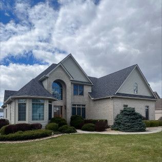 Large suburban brick house with gray roof, manicured shrubs, and cloudy sky
