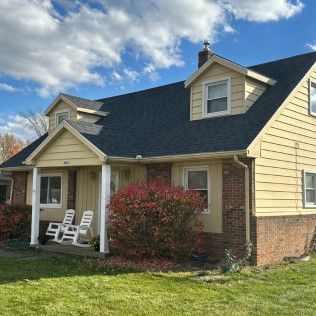 Yellow house with dark roof, front porch, and white rocking chairs on a sunny lawn
