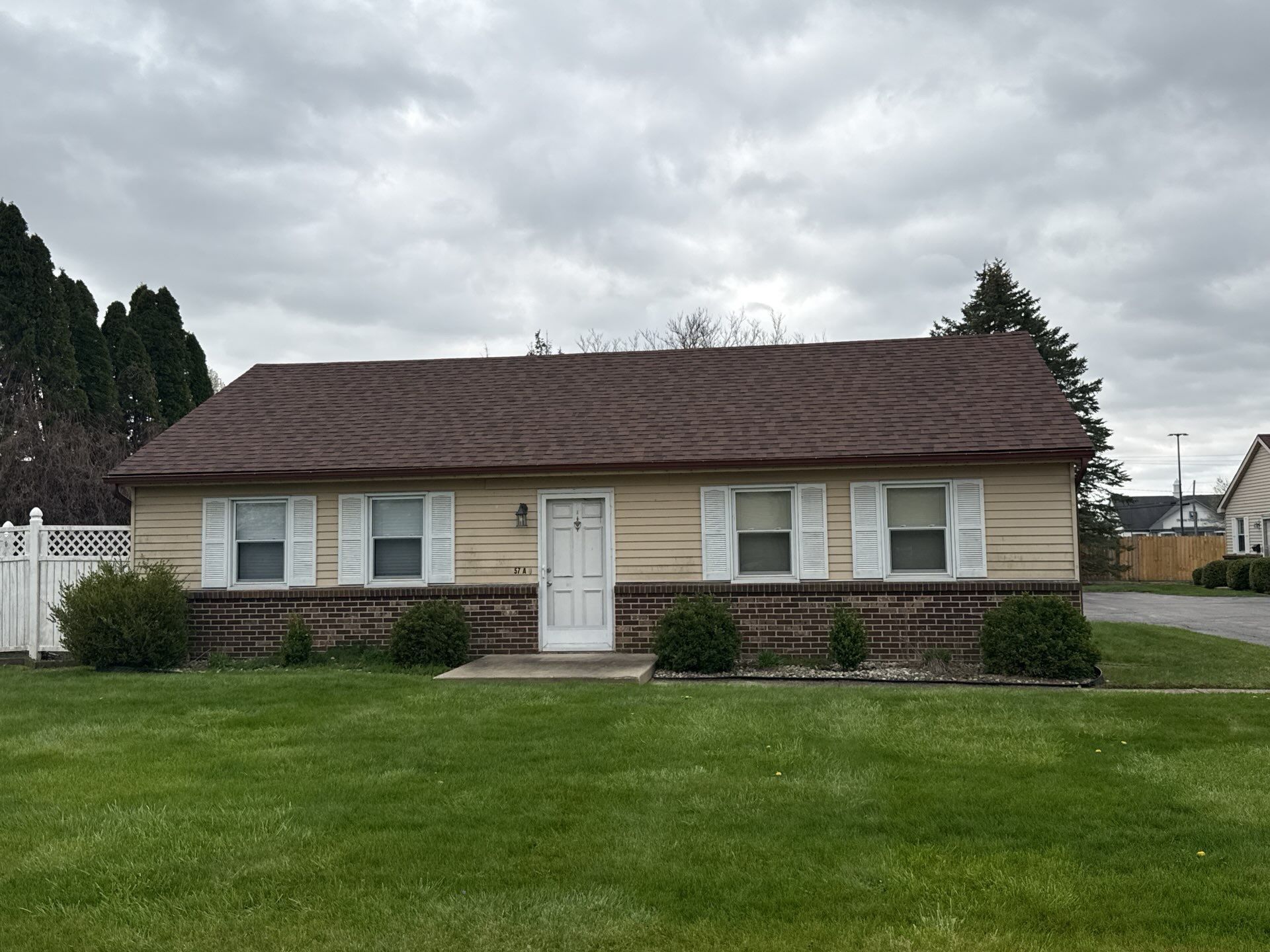 Single-story beige house with white trim, brick base, and a large green lawn under cloudy skies