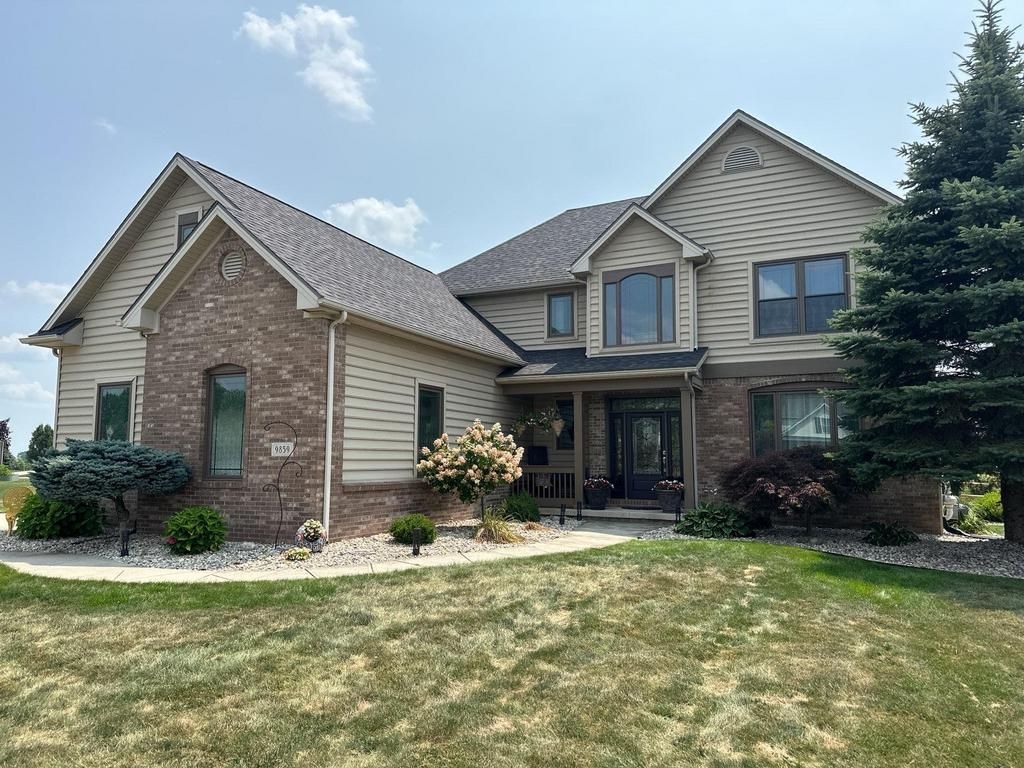 Suburban two-story house with brick and beige siding, front lawn, and trees under a blue sky