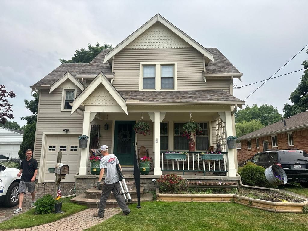 Two-story beige house with front porch; two people stand in the yard near steps and parked cars.
