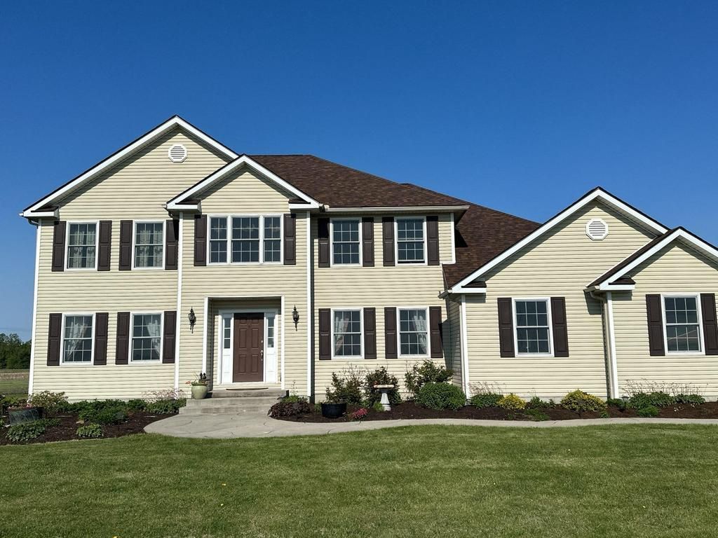 Two-story beige suburban house with black shutters and a manicured lawn under a clear blue sky