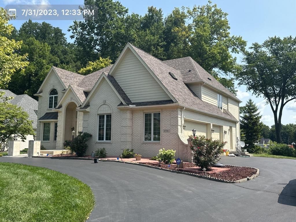 Suburban house with light siding, dark roof, and landscaped driveway on a sunny day
