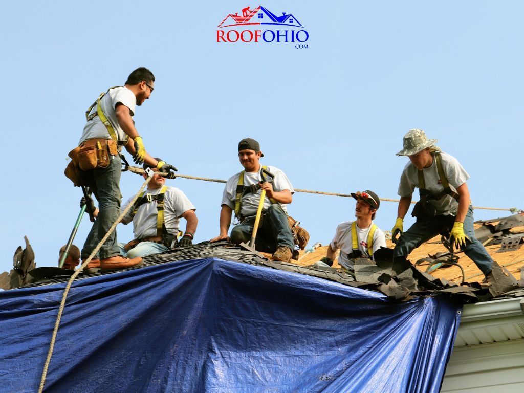 Workers on a roof pulling back a blue tarp during roof repair under a clear sky