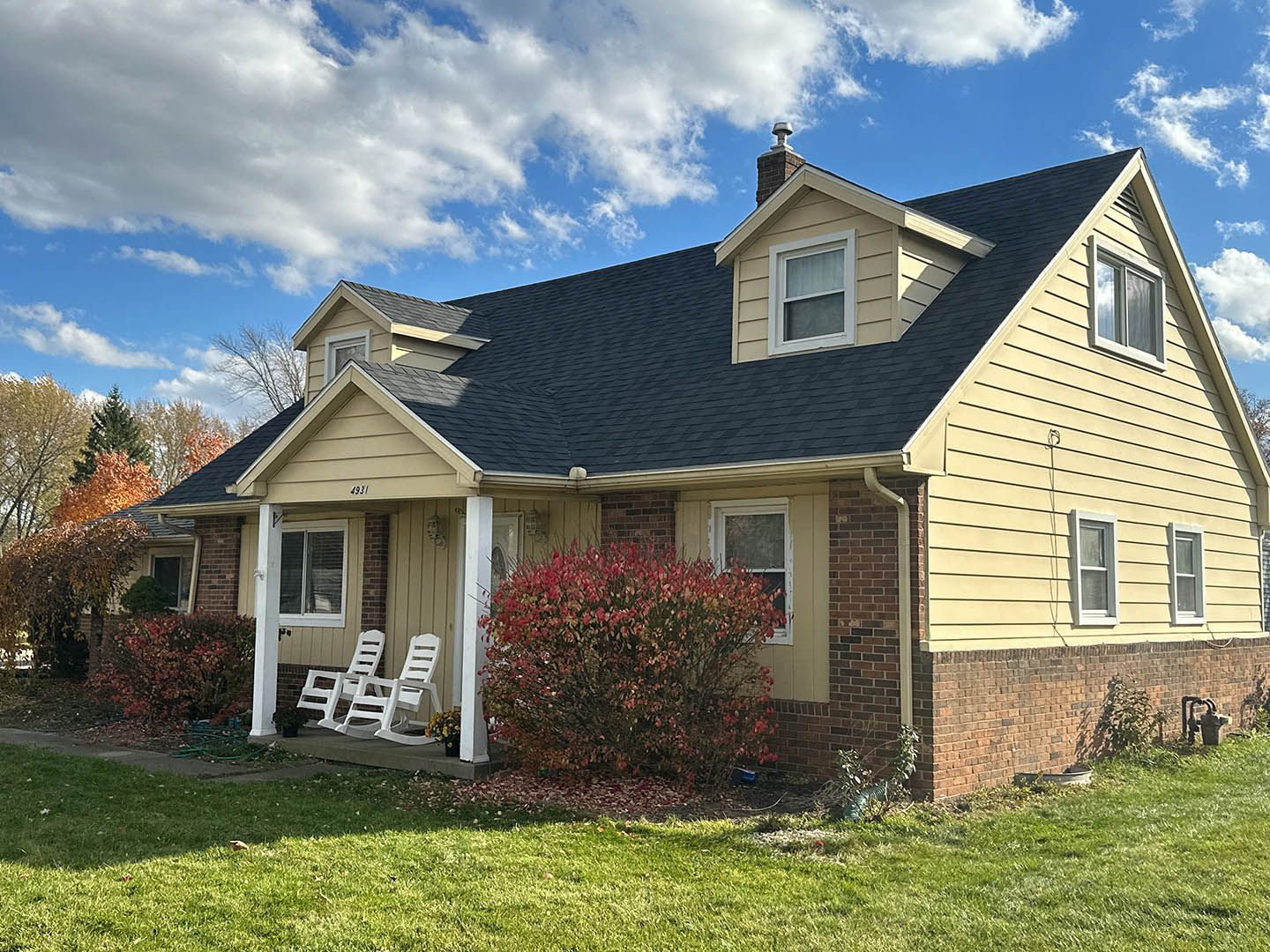 Suburban house with a front porch, white chairs, and a blue shingled roof under a partly cloudy sky