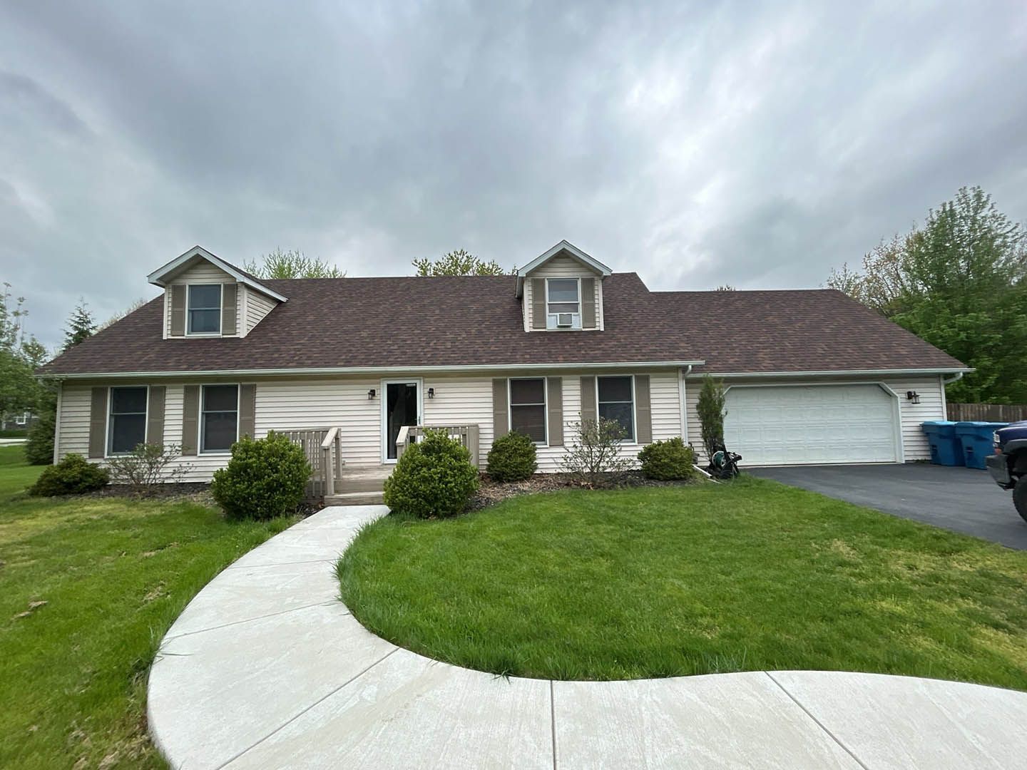 Suburban ranch house with white siding, dormer windows, curved sidewalk, and green lawn under cloudy sky
